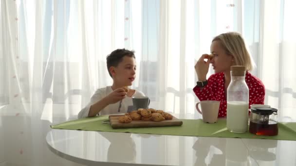 Un adorable chico caucásico lindo está comiendo galletas junto a su joven madre con el pelo rubio. El hermanito está robando galletas. . — Vídeos de Stock