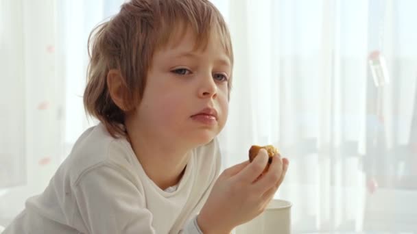 少年は、モダンなキッチンで朝食を持っています。少年は、ミルクとクッキーを食べる。キッチンの白いテーブル。テーブルの上のボウル. — ストック動画