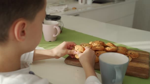 Garçon prendre le petit déjeuner dans la cuisine moderne. Garçon manger un cookie avec du lait. Table blanche dans la cuisine. Bol gris sur la table. Gros plan — Video