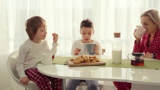 Два хлопці снідають на сучасній кухні. Хлопчик їсть печиво з молоком. Білий стіл на кухні. Мати і двоє синів снідають. Печиво з молоком . — стокове відео