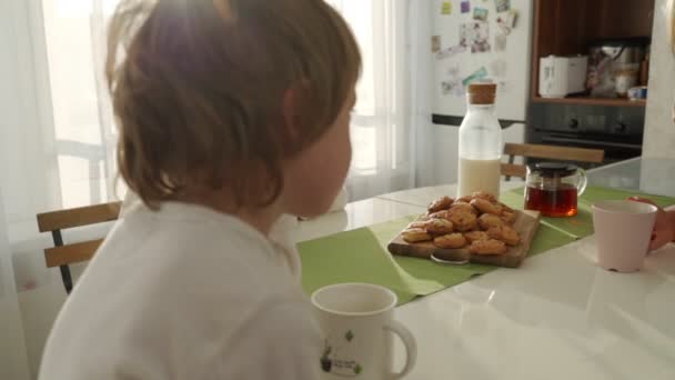 Två pojkar med frukost i moderna kök. Pojken äter kaka med mjölk. Vitt bord i köket. Mor och två söner har frukost. Cookies med mjölk. — Stockvideo