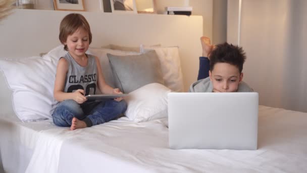 Los niños en la cama de los padres por la mañana con ordenador portátil y tableta. Los hermanos juegan juegos de ordenador. Hermanos y artilugios . — Vídeos de Stock