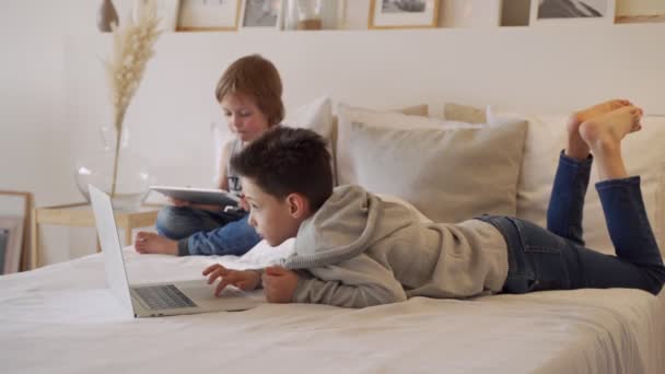 Los niños en la cama de los padres por la mañana con ordenador portátil y tableta. Los hermanos juegan juegos de ordenador. Hermanos y artilugios . — Vídeos de Stock