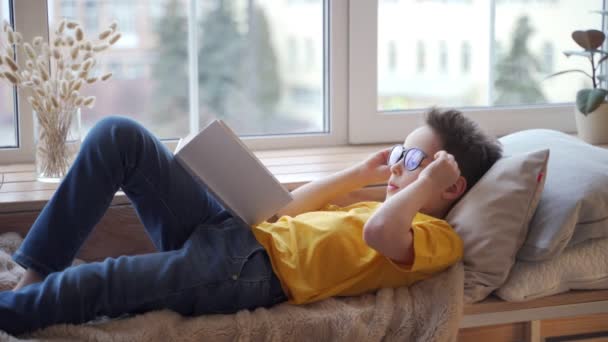 Lindo niño mintiendo en el alféizar de la ventana y leyendo libro — Vídeo de stock