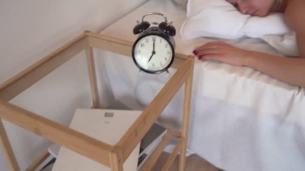 Young beautiful woman hates waking up early in the morning. Sleepy girl looking at alarm clock and trying to hide under the pillow — Stock Video