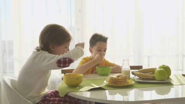 Мальчик наливает молоко в тарелку с кукурузными хлопьями, два брата завтракают — стоковое видео