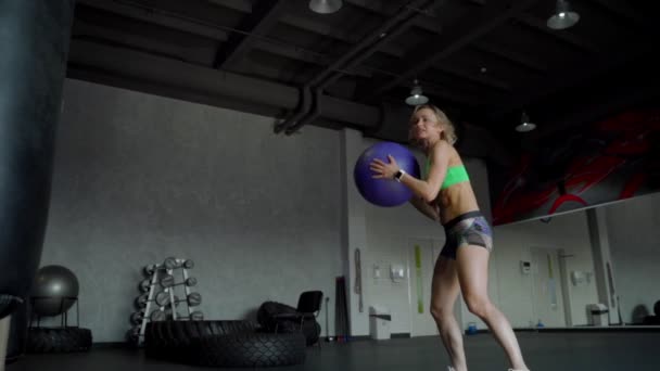 Mujer joven y fuerte con un cuerpo de fitness perfecto en ropa deportiva haciendo ejercicio con balón de medicina en el gimnasio. Chica haciendo entrenamiento crossfit. En cámara lenta. De cerca. — Vídeo de stock