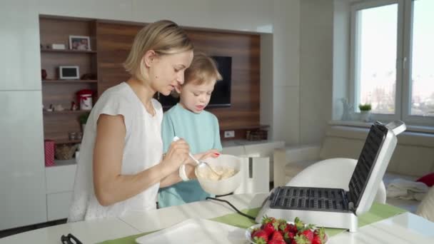 Jonge vrouw koken wafels in de keuken en schattige jongen helpt haar. Famaly koken ontbijt vroeg in de ochtend — Stockvideo