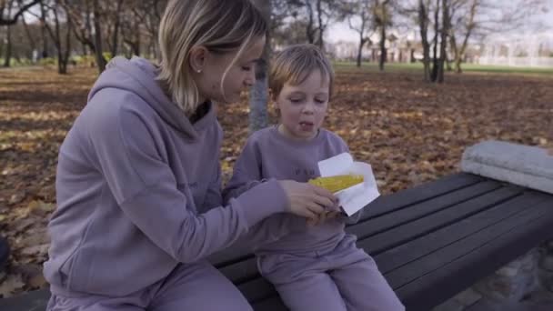 Jovem mãe com seu filho comendo milho cozido no parque no outono. Uma mulher atraente e menino estão relaxando ao ar livre — Vídeo de Stock