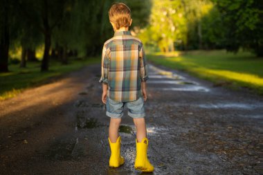 Sarı çizme giyen anaokulu çocuğu yağmurdan sonra parkta yürüyor. Güneşli bahar ya da yaz günlerinde oynayan ve eğlenen bir çocuk. Çocuklar için açık hava oyunları.