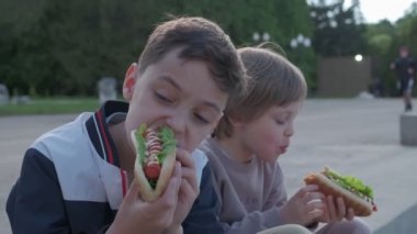 Dışarıda sosisli yiyen iki küçük çocuk. Kardeşler yemeğin tadını çıkarıyorlar. Çocuklar için sağlıksız yiyecek olarak hotdog..