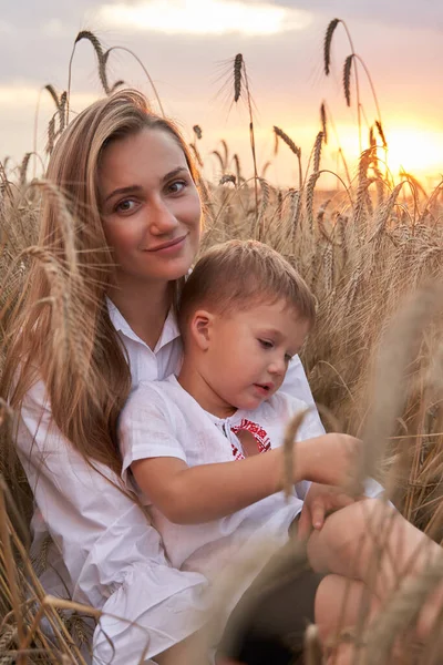 Porträtt av glad son med kamera sittandes på mödrar varv mitt i vete fält. — Stockfoto
