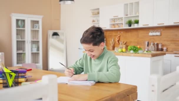 Niño en la cocina haciendo la tarea y charlando con el ordenador portátil en la mesa. — Vídeos de Stock