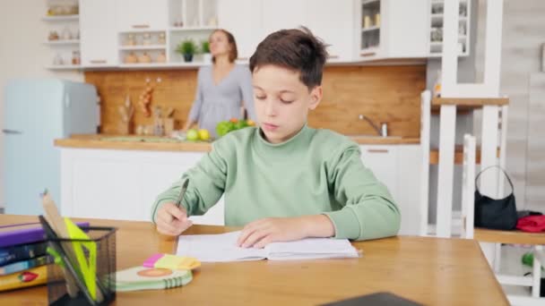 Niño en la cocina haciendo la tarea y charlando con el ordenador portátil en la mesa. — Vídeos de Stock