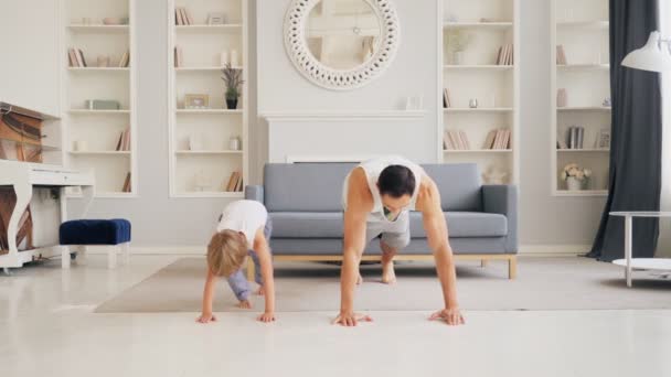 Actieve familie doet ochtendtraining in de woonkamer voor een gezonde levensstijl. Papa leert schattige kleine jongen hoe hij push-ups moet doen. Fit sportieve vader en zoon sporten thuis — Stockvideo