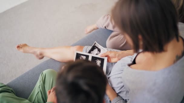 Hermosa mujer embarazada y sus hijos están sosteniendo ultrasonido escanear en su vientre en el sofá en casa, embarazada y concepto familiar — Vídeos de Stock