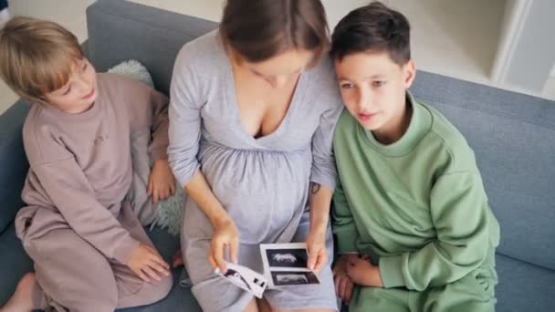 Gyönyörű terhes nő és fiai tartanak ultrahang vizsgálat a hasán kanapén otthon, terhes és családi koncepció — Stock videók
