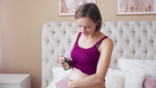 Une heureuse femme enceinte applique de l'huile hydratante des vergetures à son estomac assis à la maison sur le lit, elle caresse doucement son ventre et sourit. Le concept d'une grossesse en santé. — Video