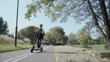 Güneşli bir günde şehirde elektrikli scooterıyla gezen okul çocuğu. Renkli elbiseli çocuk okula giderken bisiklete biniyor. Elektrik taşımacılığı