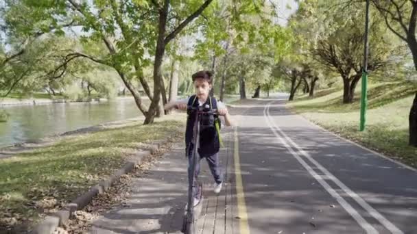 Iskolás fiú lovagol az elektromos robogójával a városban, hátizsákkal a napsütéses napon. Színes ruhás gyerek biciklizik az iskolába menet. Elektromos közlekedés — Stock videók