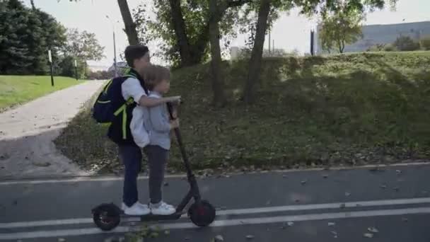 Dwóch chłopców idzie do szkoły na skuterze elektrycznym w parku z plecakiem w słoneczny dzień. Transport elektryczny — Wideo stockowe