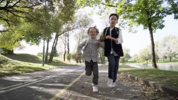 Два мальчика из начальной школы с рюкзаками бегут в школу после перерыва в парке. Мальчики бегают с рюкзаками в солнечный день. Медленное движение — стоковое видео