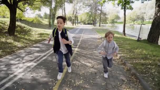 Två lågstadiekillar med ryggsäckar som springer till skolan efter rasten i parken. Pojkar springer med ryggsäckar på soliga dagar. Långsamma rörelser — Stockvideo