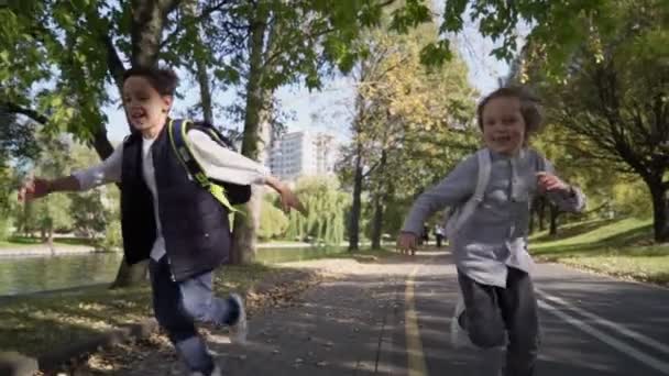 Två lågstadiekillar med ryggsäckar som springer till skolan efter rasten i parken. Pojkar springer med ryggsäckar på soliga dagar. Långsamma rörelser — Stockvideo