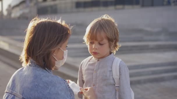 A mãe escolta o filho para a escola. Uma mãe coloca uma máscara protetora para seu filho antes de entrar na escola. O início do ano letivo — Vídeo de Stock