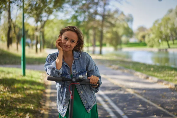 Mujer feliz montando en un e-scooter. Joven y hermosa mujer caucásica monta scooter eléctrico en el parque —  Fotos de Stock