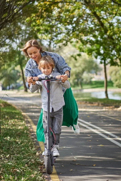 Happy woman riding on an e-scooter with her son. Young and beautiful caucasian woman rides electric scooter with boy in park Royalty Free Stock Images