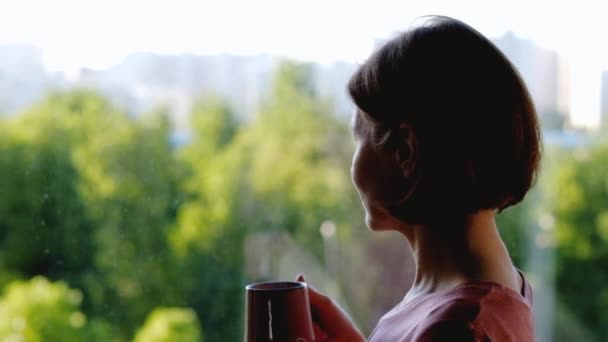 Frau steht am Fenster und trinkt Kaffee oder Tee — Stockvideo