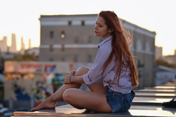 Όμορφη γυναίκα με τα κόκκινα μαλλιά που κάθεται σε μια ταράτσα ή γέφυρα, το φως του ηλιοβασιλέματος — Φωτογραφία Αρχείου