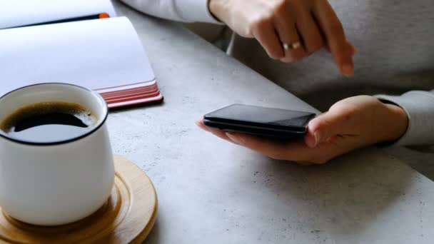 Смартфон в женских руках, кофе и блокнот на столе. Закрыть — стоковое видео