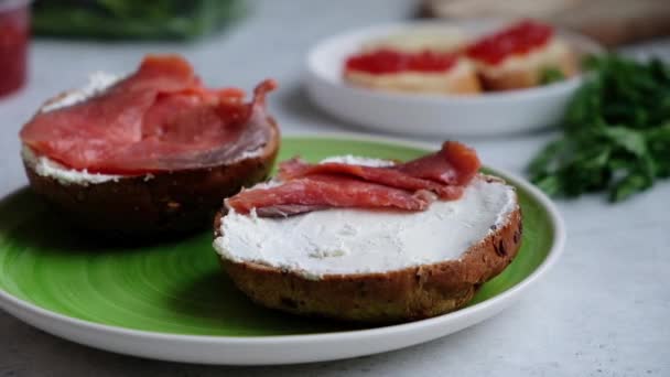 Uma mão de chefs coloca fatias de salmão vermelho em um pão escuro de gergelim com queijo creme, em um prato verde em luz suave. . — Vídeo de Stock