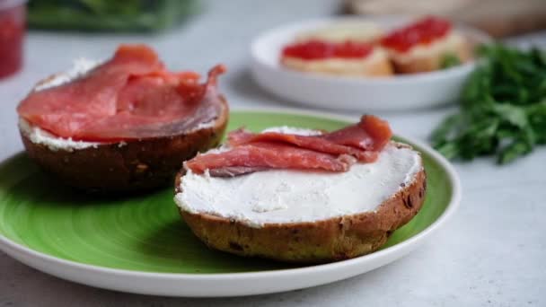 En kockar hand släpper skivor av röd lax på en sesam mörkt bröd med färskost, på en grön platta i mjukt ljus. — Stockvideo