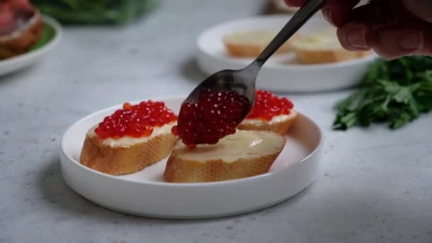 Uma mão de mulher coloca caviar vermelho em uma baguete com manteiga, em uma placa branca em luz suave. . — Vídeo de Stock