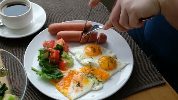 Un hombre desayunando. Cuchillo y tenedor en la mano. Desayuno americano con huevos fritos, salchichas, ensalada y tostadas . — Vídeos de Stock