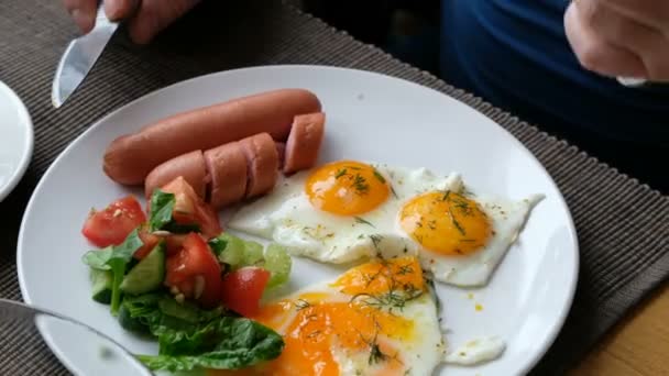 Um homem a tomar o pequeno almoço. Faca e garfo na mão. Café da manhã estilo americano com ovos fritos, salsicha, salada e torrada . — Vídeo de Stock