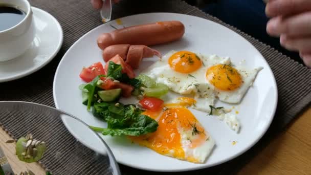 Ο άνθρωπος τρώει πρωινό. Μαχαίρι και πιρούνι στο χέρι. Αμερικάνικο πρωινό με τηγανητά αυγά, λουκάνικο, σαλάτα και τοστ. — Αρχείο Βίντεο