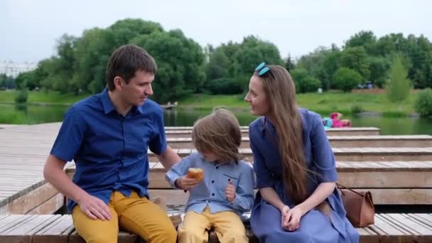 Happy familj Concept: mor, far och liten pojke som sitter vid floden och äter croissant — Stockvideo