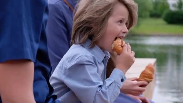 Kisfiú ül a folyó a szüleivel, és étkezési croissant — Stock videók