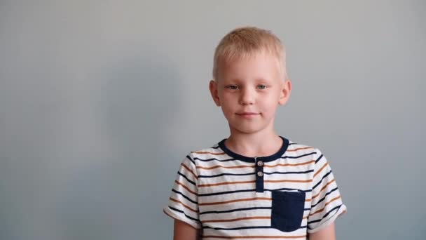 Χαριτωμένο αγόρι 6 χρονών παίζοντας κοιτάζοντας την κάμερα και χαμογελώντας. Γκρι φόντο — Αρχείο Βίντεο