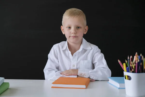 Je retourne à l'école. Mignon petit garçon assis à la table sur le tableau noir backgrpund. Enfant de l'école primaire. Concept d'éducation . — Photo