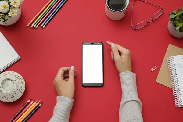 Kırmızı ofis masasında beyaz cep telefonu tutan ellerin mockup görüntü. Üst görünüm, düz yatıyordu.