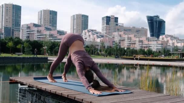 Młoda kobieta robi ćwiczenia jogi z miastem na tle. Adho Mukha shvanasana — Wideo stockowe
