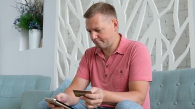 Genç beyaz adam oturma odasında kredi kartı ile online alışveriş için akıllı telefon kullanarak. .