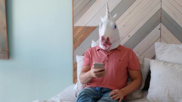 Strano video divertente: l'uomo in una maschera di unicorno sdraiato su un letto e guardando smartphone — Video Stock