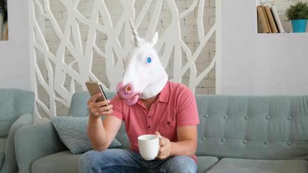 Extraño vídeo divertido: hombre en una máscara de unicornio sentado en un sofá y sosteniendo un teléfono inteligente y beber café — Vídeo de stock