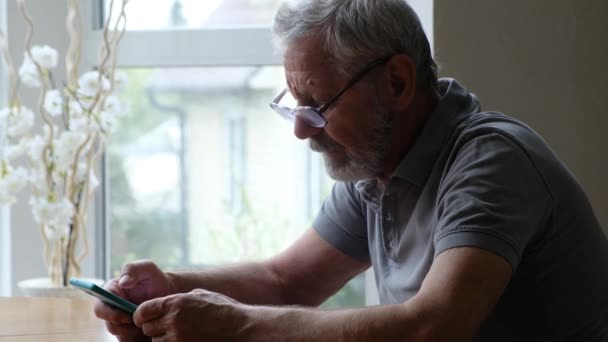 Senior man i glasögon sitter vid bordet och tittar på mobiltelefon hemma — Stockvideo